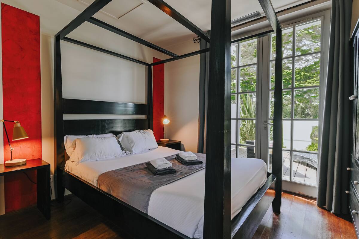 Un lit à baldaquin avec du linge de lit blanc, une table de chevet avec une lampe et une vue sur la verdure à travers de grandes fenêtres.