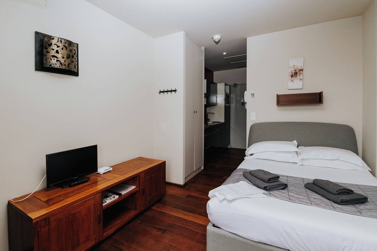 Une chambre d’hôtel moderne avec un meuble télé en bois, un lit où les serviettes sont soigneusement pliées, et un couloir menant à un coin cuisine.