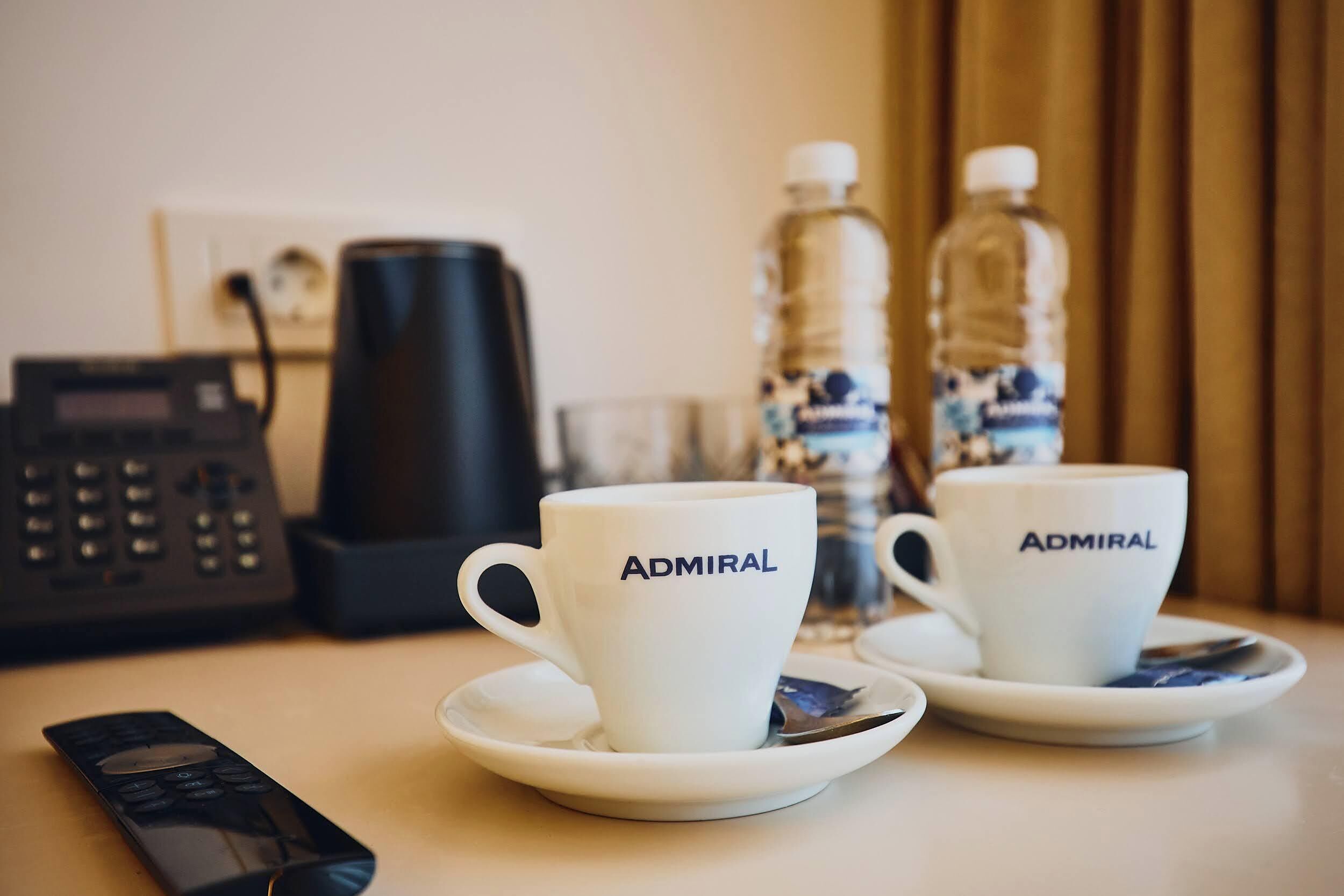 Dos tazas blancas con la palabra "Admiral", un control remoto, una cafetera y dos botellas de agua sobre una mesa.