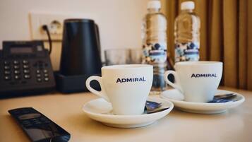 Two white cups with the word "Admiral" on them, a remote control, a coffee maker, and two water bottles on a table.
