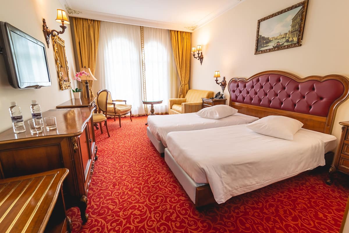 Habitación de hotel con dos camas, un escritorio con lámpara, una silla, un televisor y un cuadro en la pared.