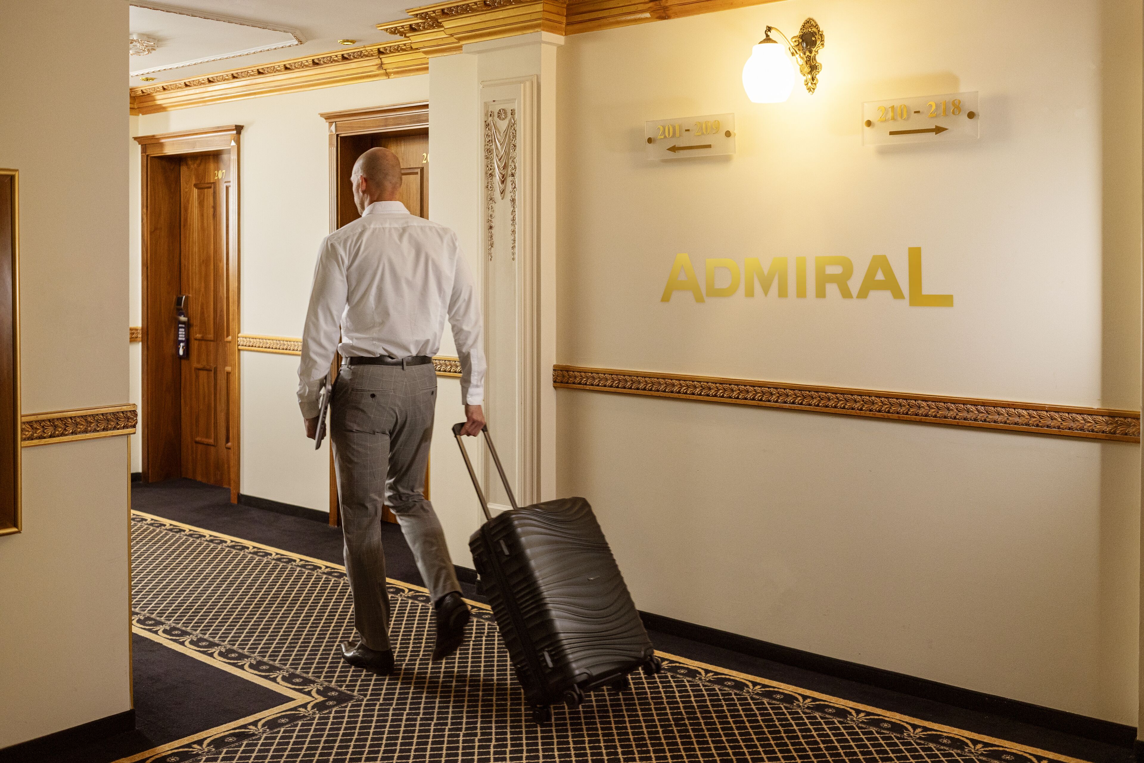 Un uomo cammina lungo un corridoio trascinando una valigia, diretto verso una porta con la scritta "ADMIRAL".