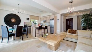 A spacious living room with a large sofa, a dining area with chairs, a kitchen with a refrigerator, and a chandelier.