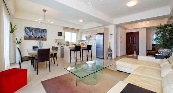 A modern living room with a glass coffee table, a dining area with chairs, a kitchen with a refrigerator, and a red accent chair.
