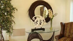 A round mirror on a glass table with a decorative white sculpture, a vase with flowers, and a patterned armchair.