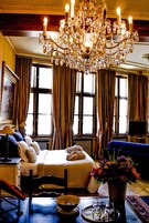 Una habitación con una lámpara de araña grande, una cama con almohadas, un sofá azul, un jarrón con flores y una bandeja con saleros y pimenteros.