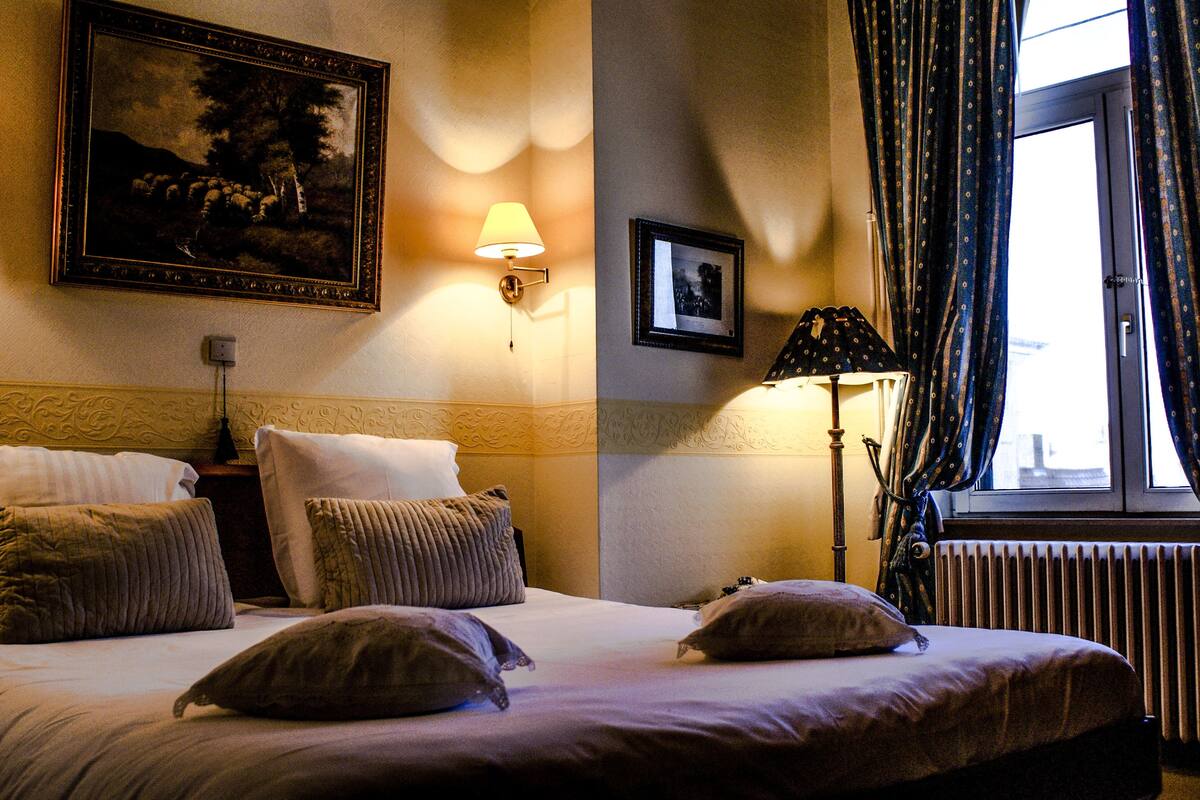 A hotel room with a bed, pillows, a lamp, a framed picture, and a window with curtains.