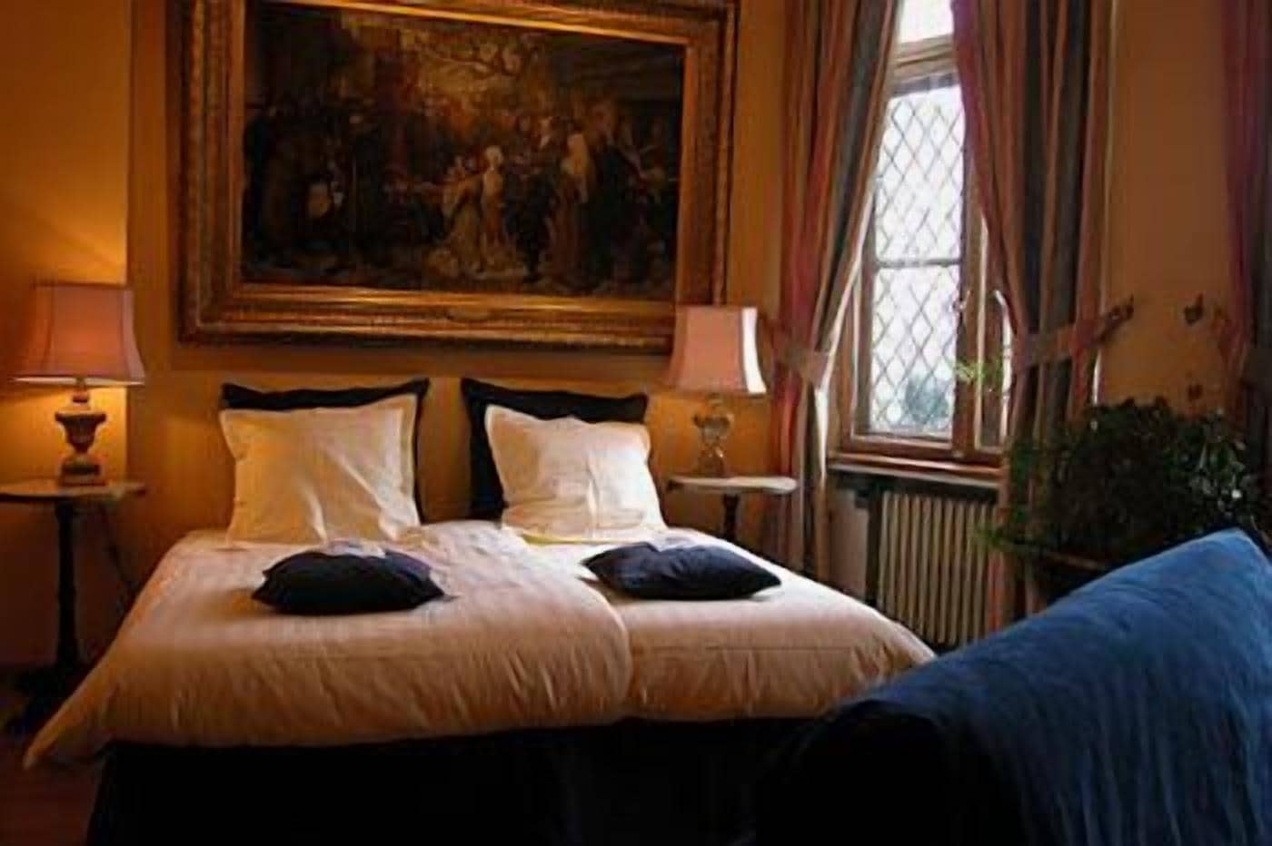A hotel room with a large bed, two bedside tables with lamps, a window with curtains, and a painting on the wall.