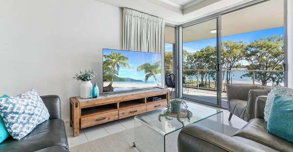 A modern living room with a large TV, a glass coffee table, and a sofa. - On the Bay Apartments (Bongaree)
