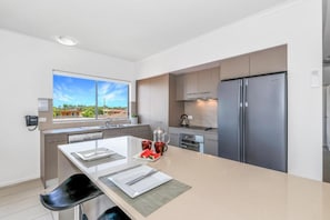 A modern kitchen with a large island, stainless steel appliances, and a view of a residential area. - On the Bay Apartments (Bongaree)
