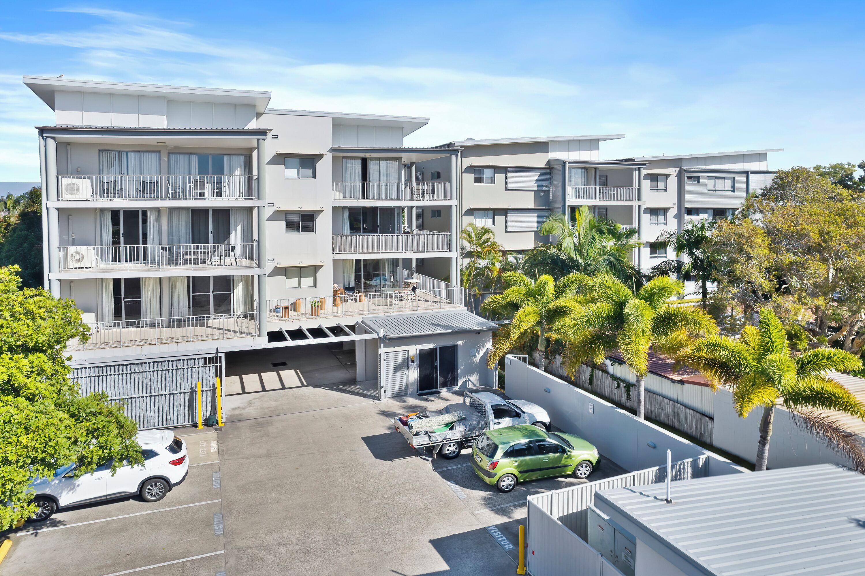 A multi-story residential building with balconies, a parking area, and palm trees.