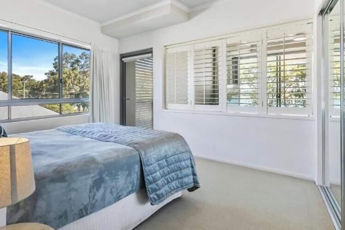 A bedroom with a bed, a lamp, a window with blinds, and a view of trees.