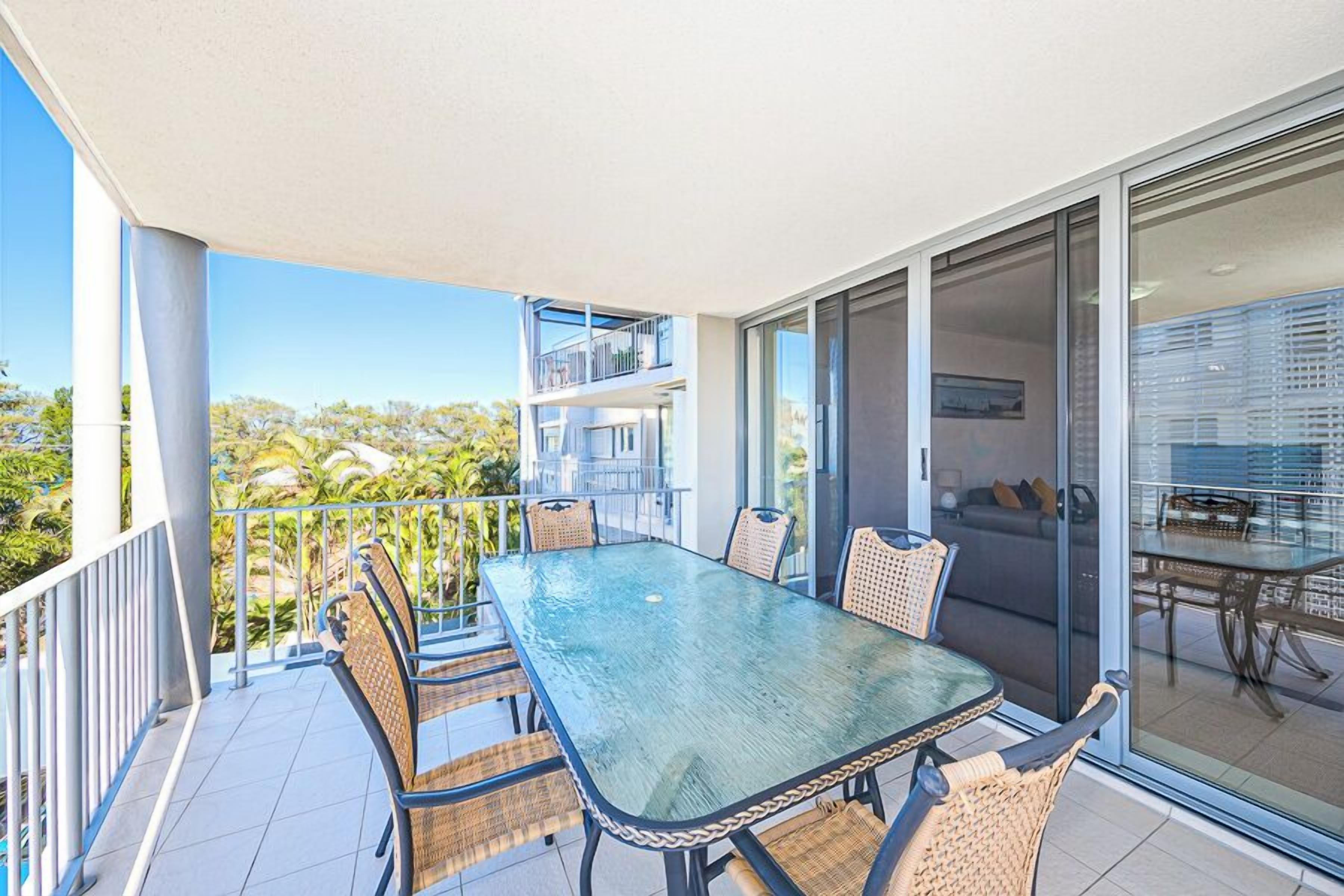 A balcony with a glass table and wicker chairs, overlooking a residential area with houses and greenery.