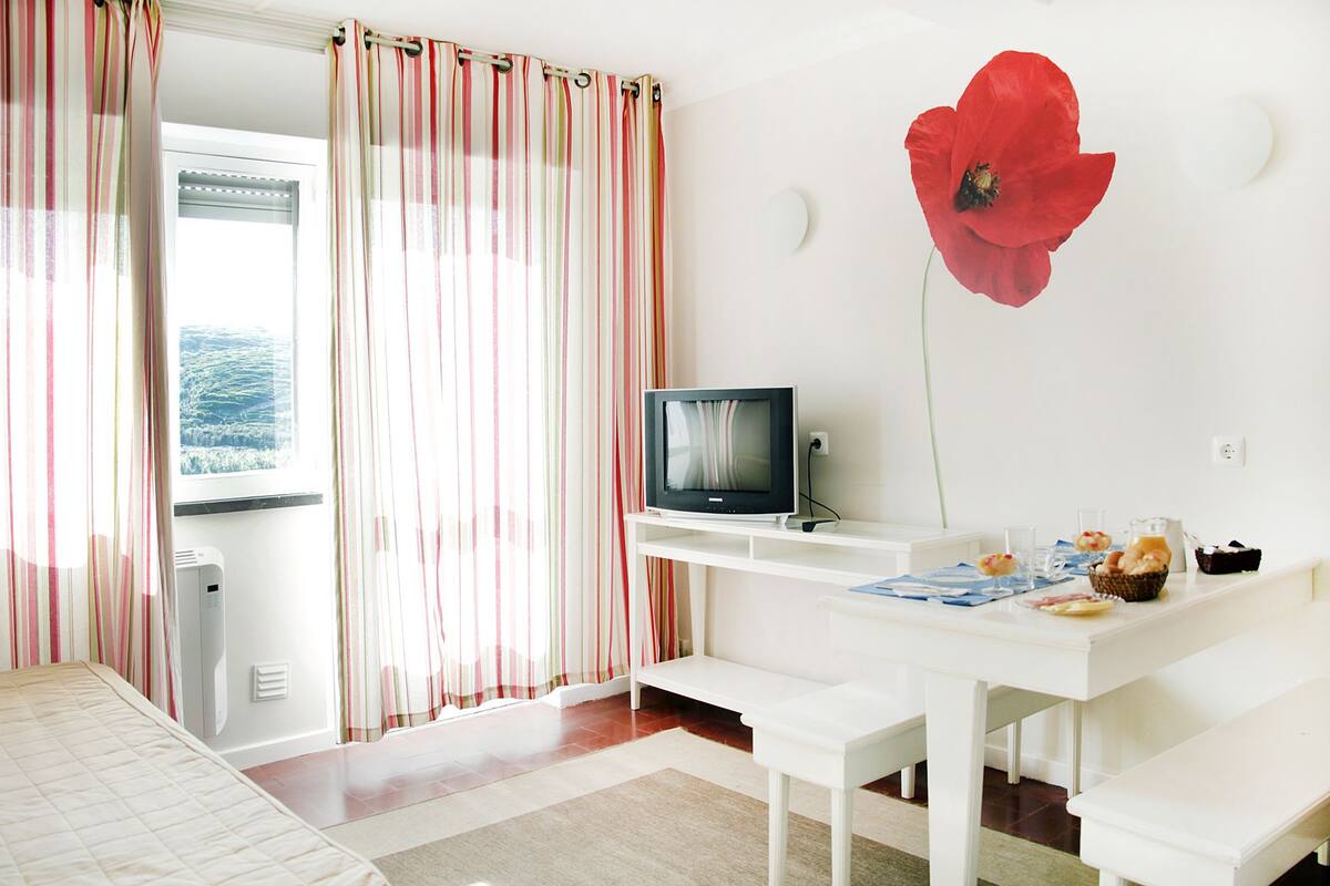 A room with a bed, a TV on a white stand, a poppy flower decoration, and a table with food and drinks.