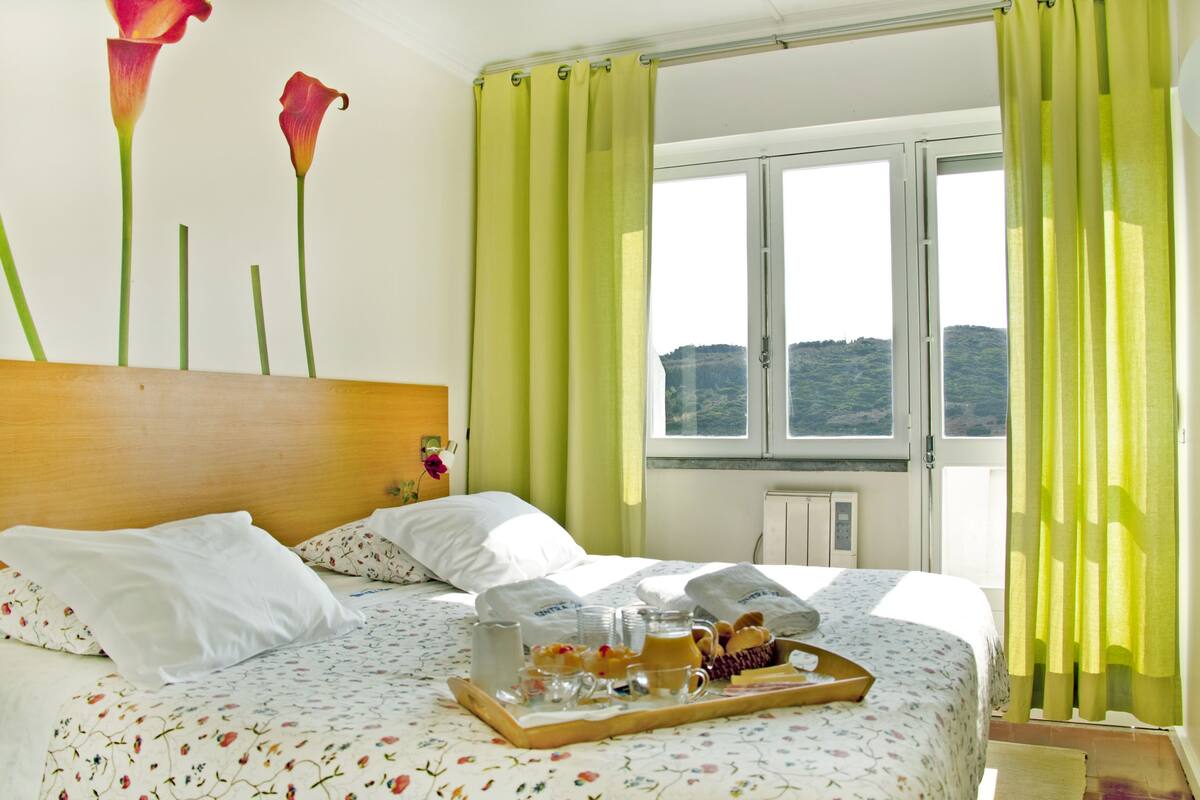 A neatly made bed with floral bedding, a tray of breakfast items, and a view of a mountainous landscape through the window.