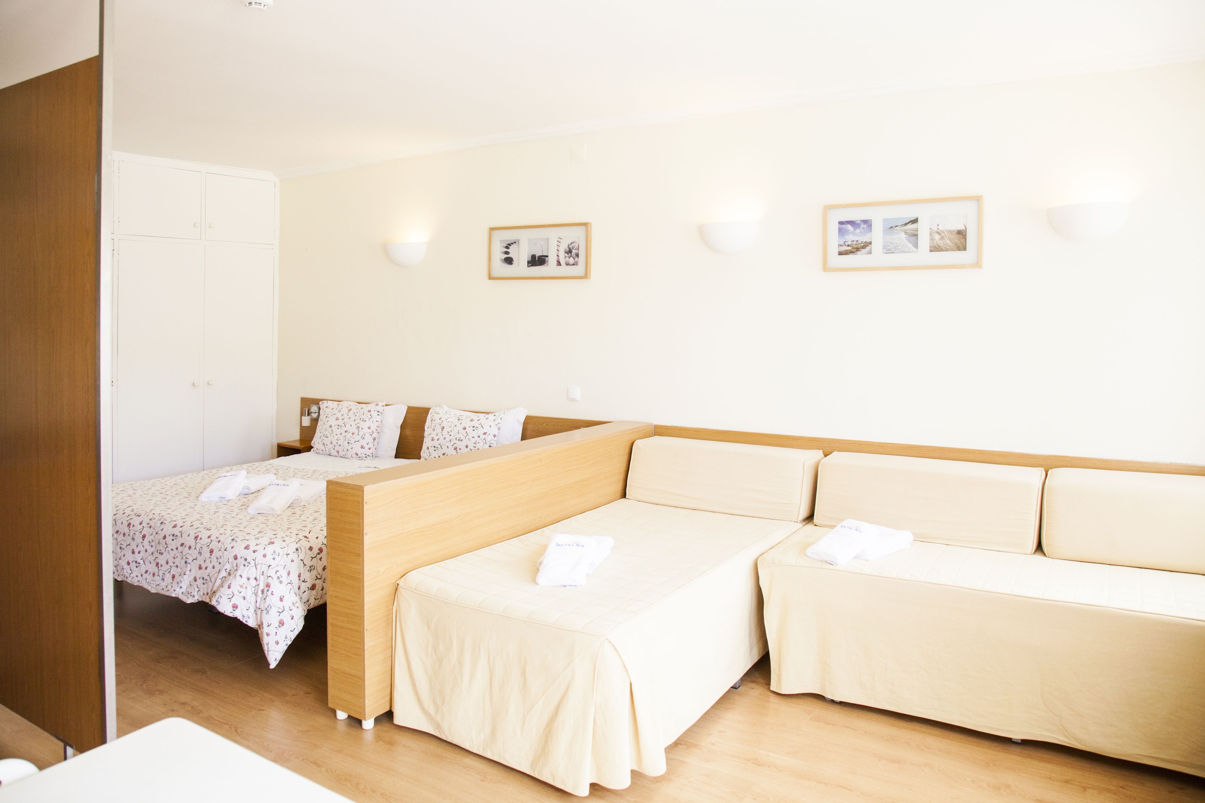 A hotel room with a bed, a sofa, and a small table. There are two wall-mounted light fixtures and two framed pictures on the wall.