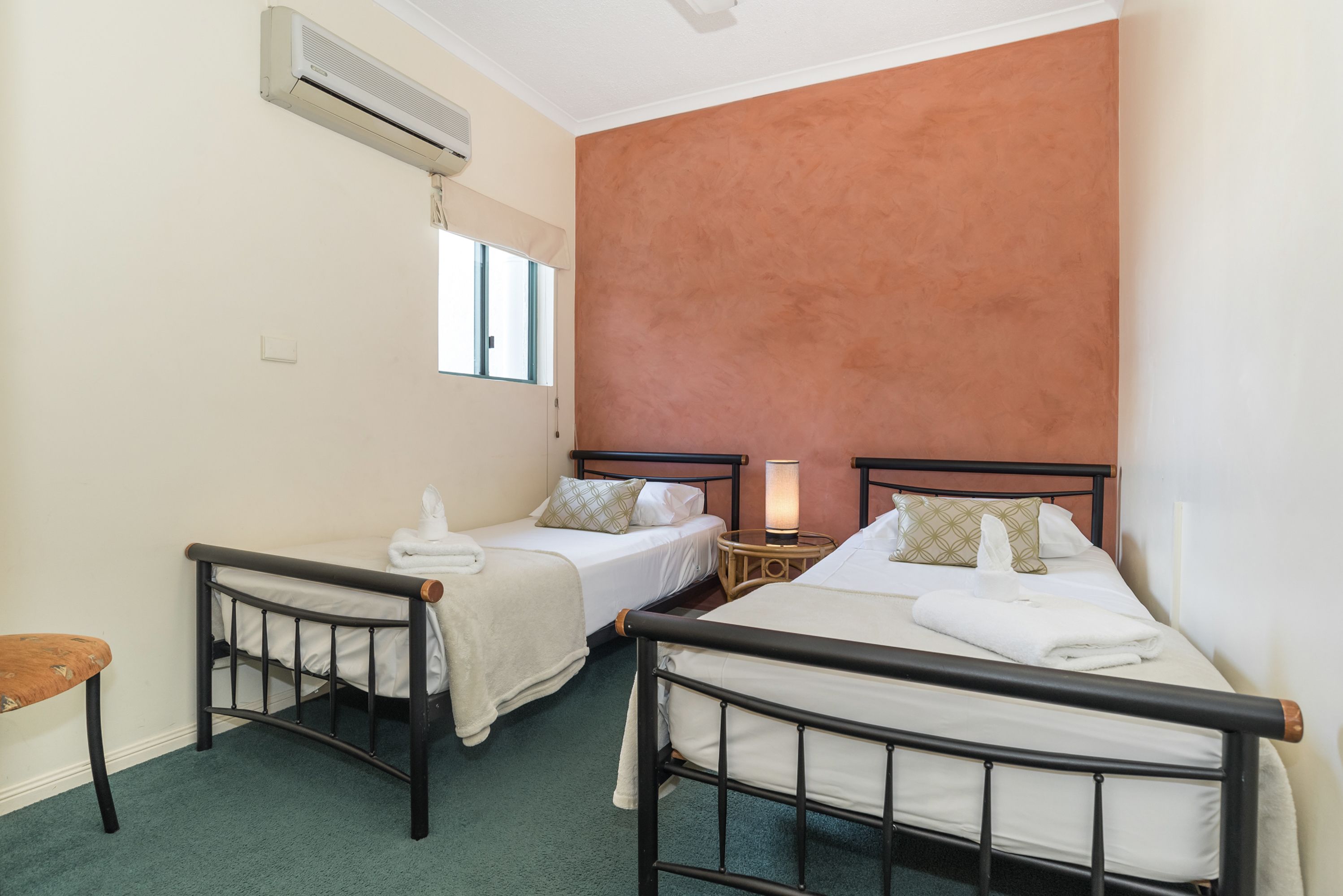 A hotel room with two single beds, a small table with a lamp, a chair, and an air conditioning unit.