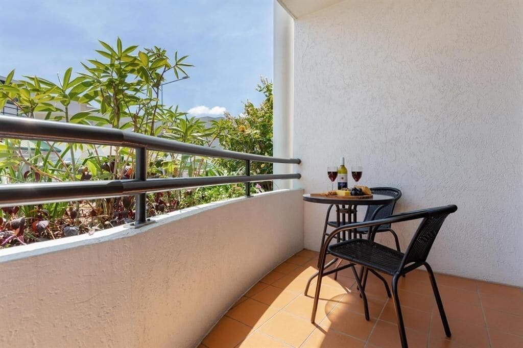 A small balcony with a table and chairs, a railing, and a view of greenery.