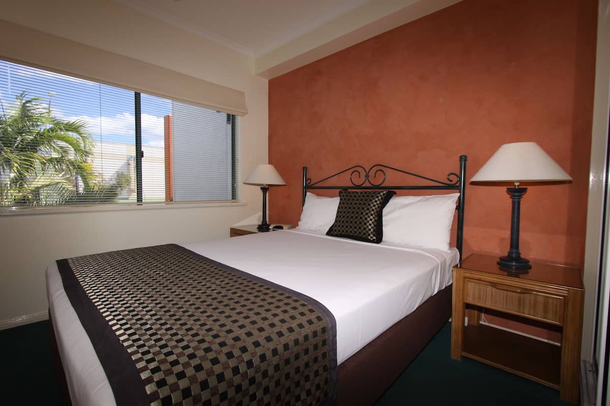 Habitación de hotel con una cama grande, dos mesitas de noche, una ventana con persianas y vista a palmeras.