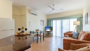 A modern living room with a flat-screen TV, a sofa, a dining table with chairs, and a kitchen area with a refrigerator and wine glasses.