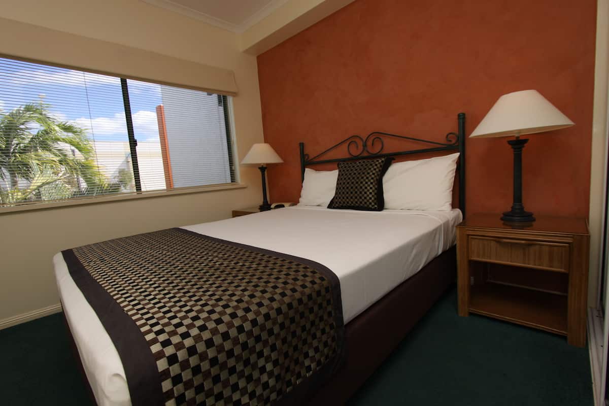 Habitación de hotel con una cama grande, dos mesitas de noche con lámparas, una ventana con persianas y vistas a palmeras.