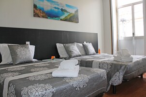 Quarto de hotel com duas camas, colchas de padrão cinzento, almofadas brancas e um quadro com paisagem costeira na parede.