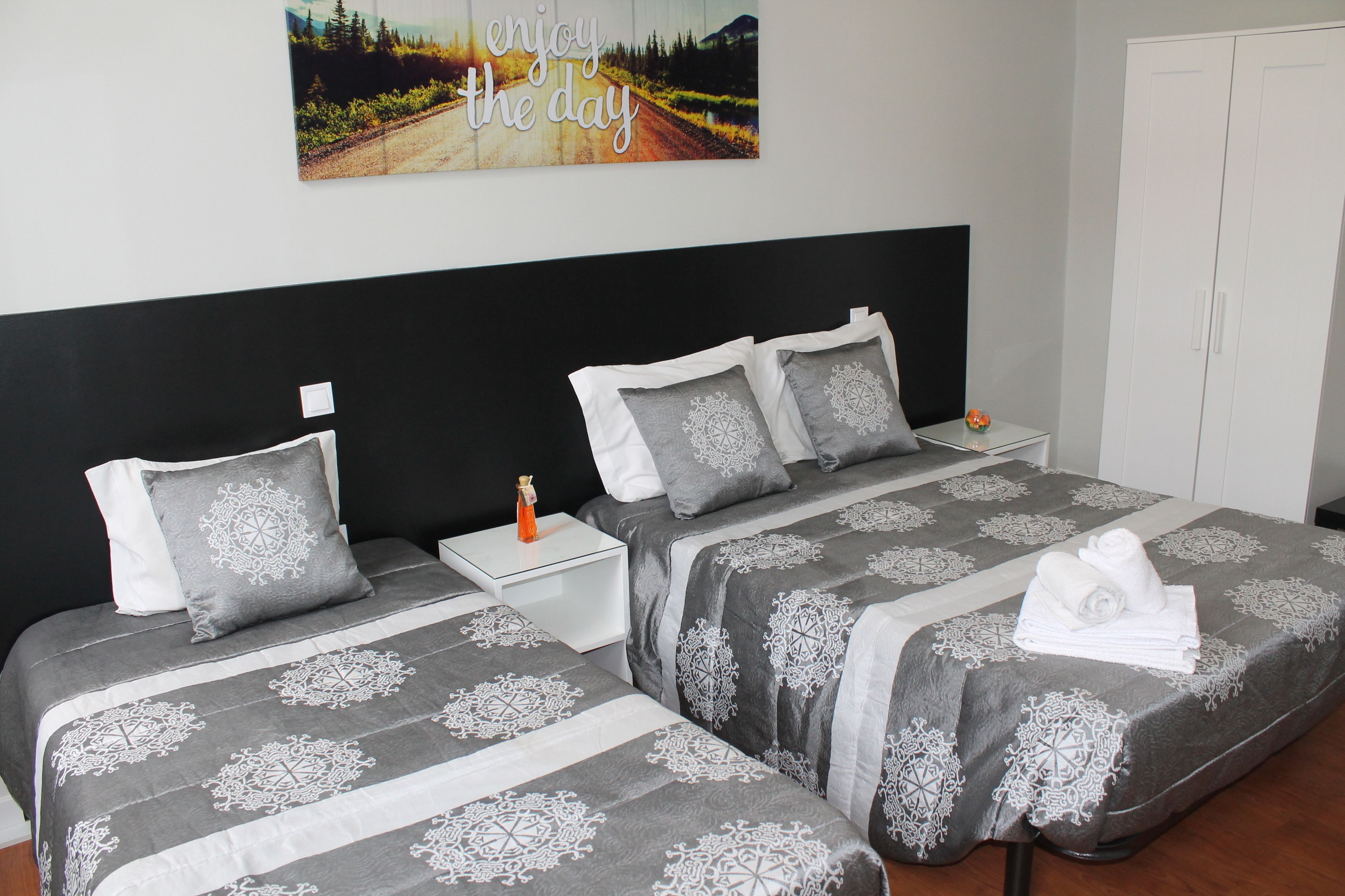 A bedroom with two beds, each with a grey and white patterned bedspread, a nightstand with a lamp, and a wall-mounted picture with the text "enjoy the day".