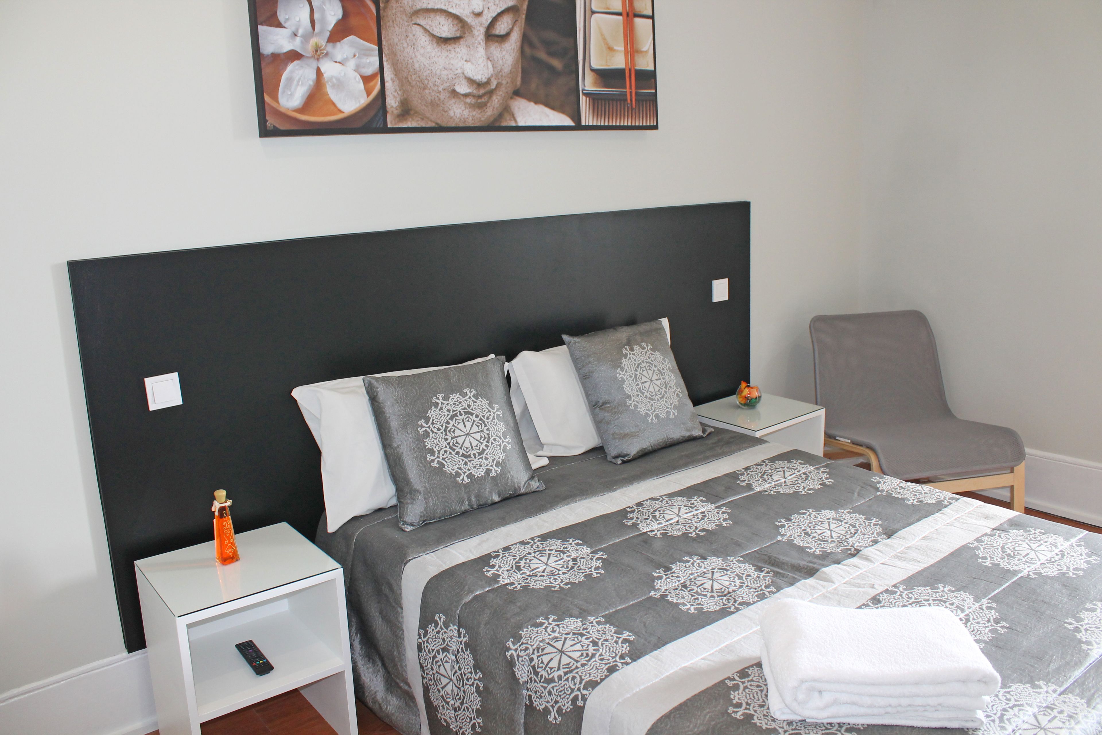 A neatly made bed with a patterned bedspread, a small bedside table with a remote control, and a chair in the corner.