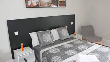 A neatly made bed with a patterned bedspread, a small bedside table with a remote control, and a chair in the corner.