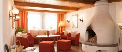 A cozy living room with wooden beams, red upholstered furniture, a fireplace, and a table with various items.