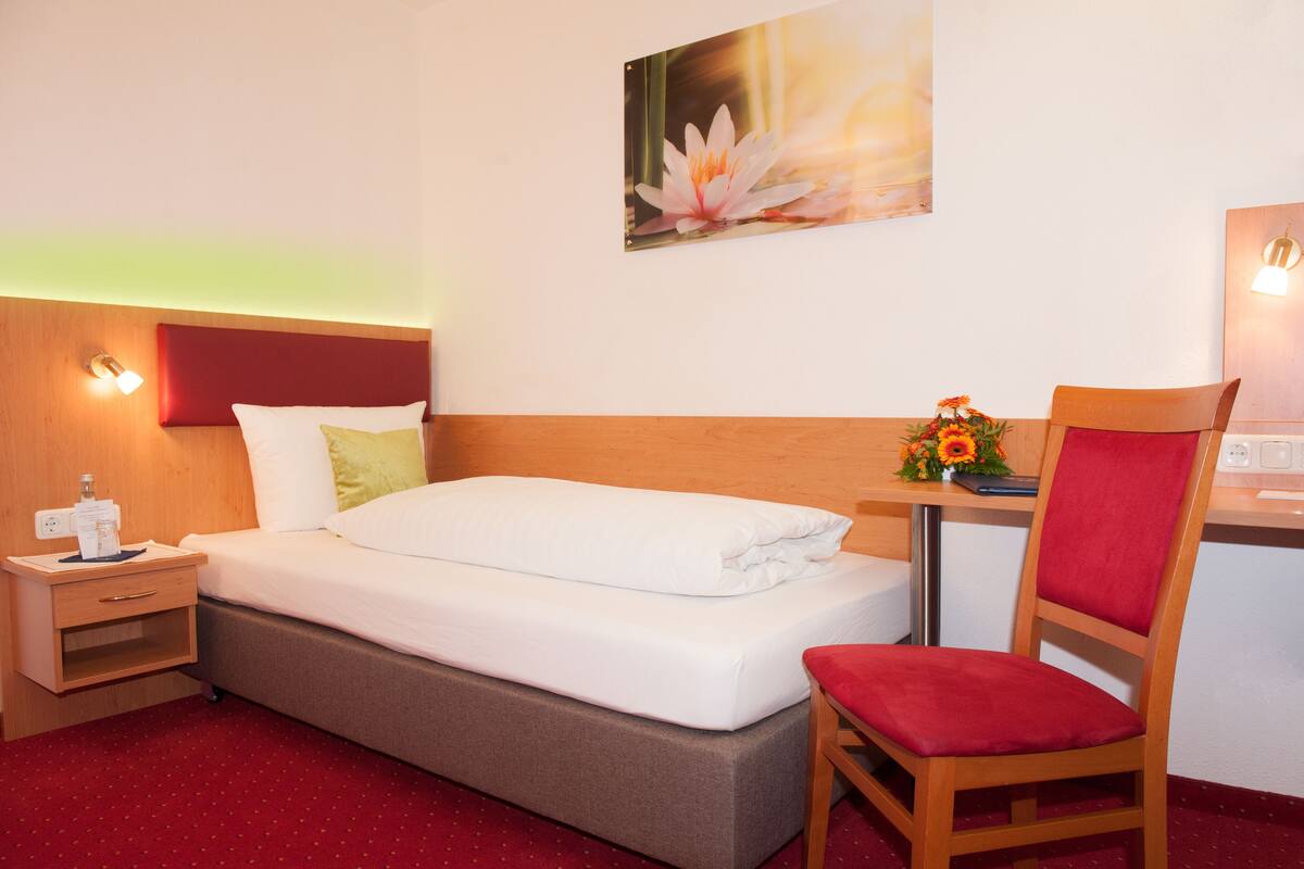 Quarto de hotel com cama, mesa de cabeceira, escrivaninha, cadeira e luminária na parede.