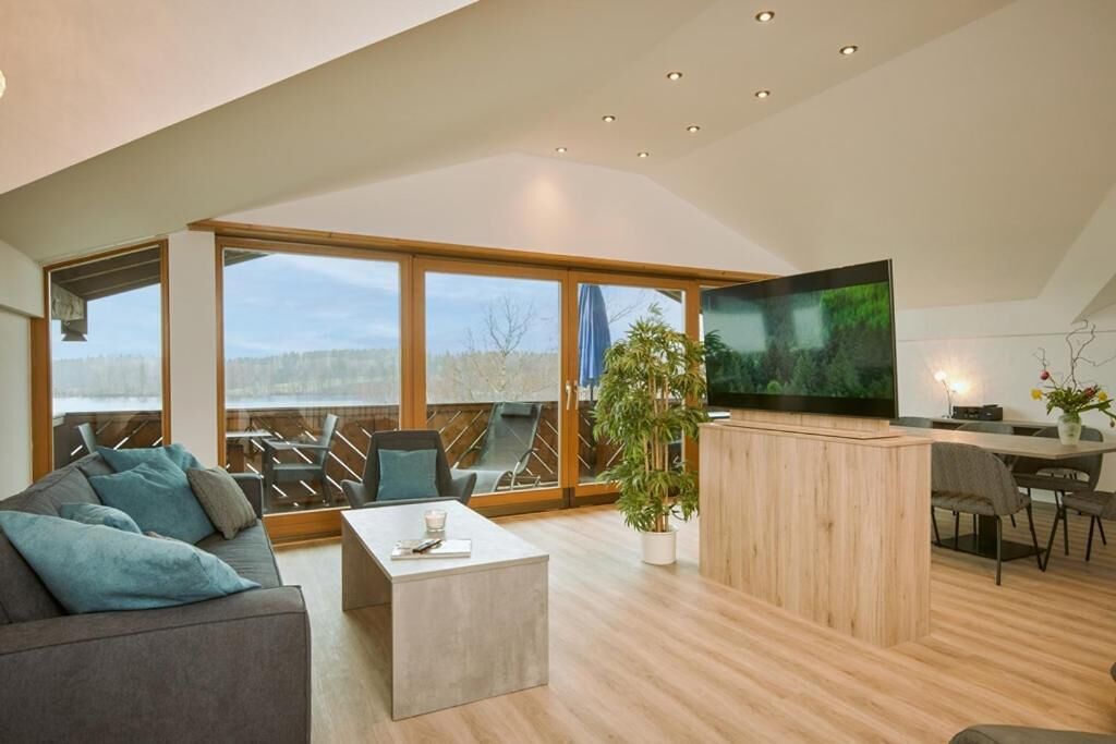 Una sala de estar moderna con un televisor grande, suelo de madera y vistas amplias a través de grandes ventanales.