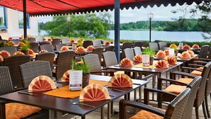 An outdoor dining area with tables set for guests, featuring wicker chairs and orange cushions, overlooking a lake. - Aktiv- und Wellnesshotel Seeblick (Bad Endorf)