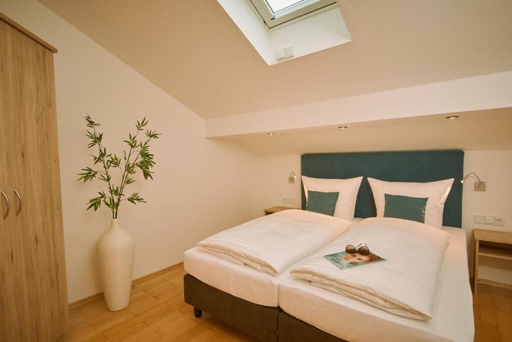 A bedroom with a bed, a nightstand, a bedside table, a lamp, a window, and a bamboo plant.