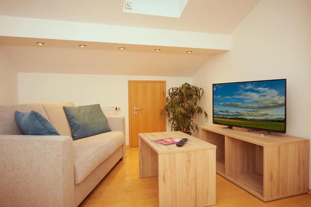 Una sala de estar moderna con un sofá, una mesa de centro, un televisor sobre un mueble de madera y una puerta al fondo.