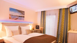 A hotel room with a large bed, a desk, a chair, a TV, and a window with curtains. - Aktiv- und Wellnesshotel Seeblick (Bad Endorf)