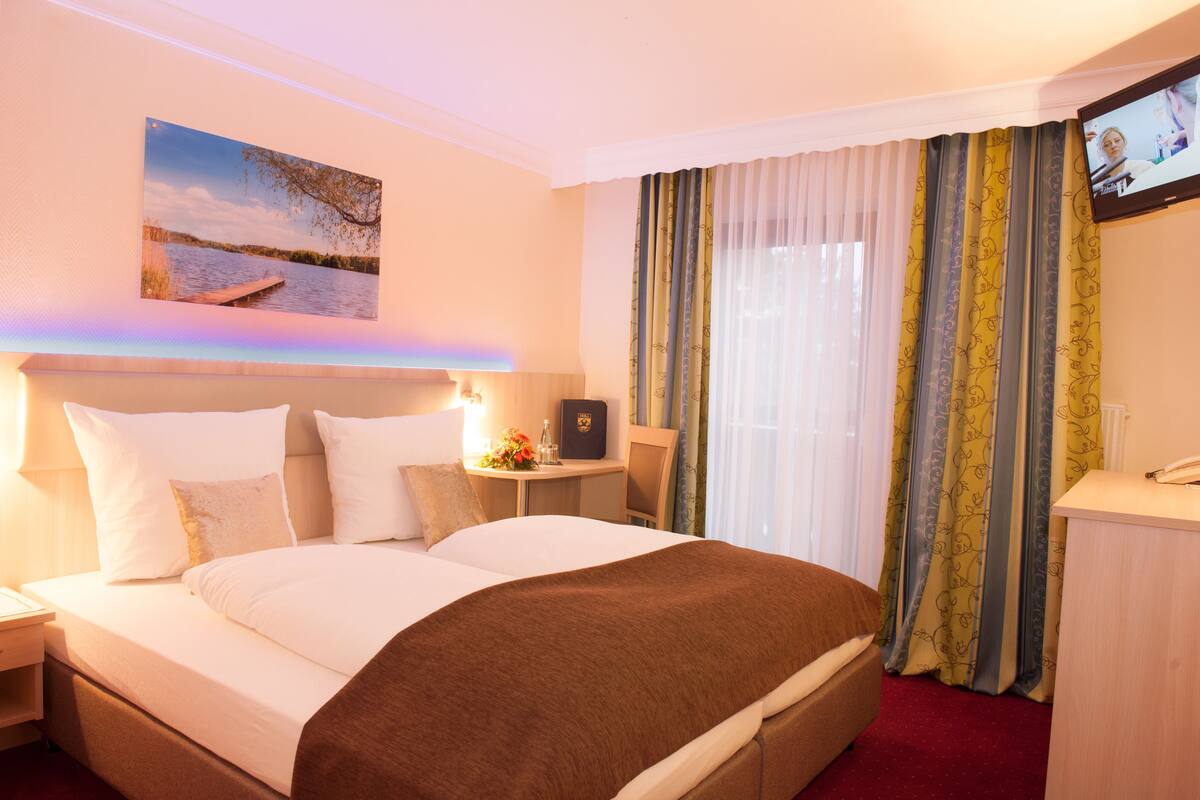 Quarto de hotel com uma cama grande, escrivaninha, cadeira, televisão e janela com cortinas.