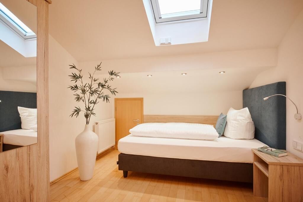 A bedroom with a skylight, a bed with white bedding, a bedside table, and a large white vase with a plant.