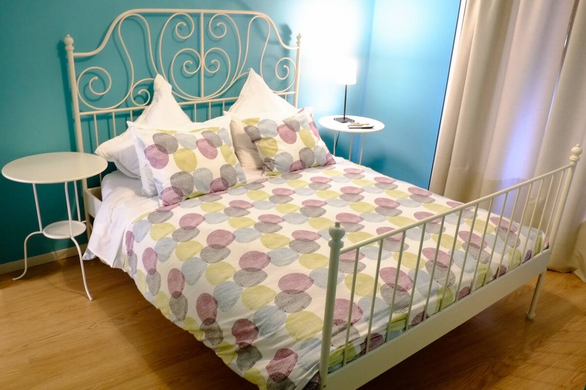 A neatly made bed with a polka dot bedspread, two white pillows, and a metal headboard. There is a small round table with a lamp beside the bed.