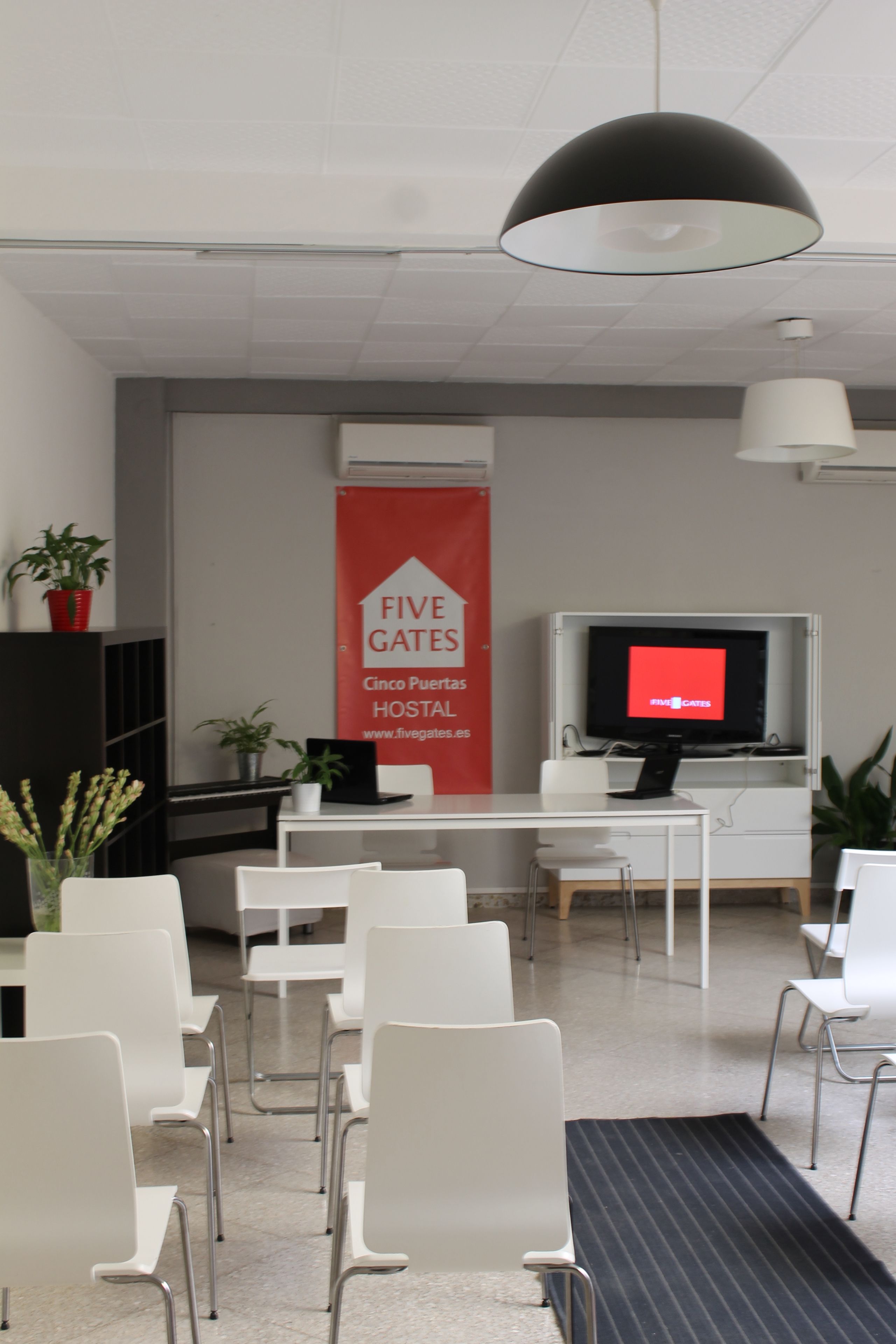 Un espace d’accueil avec une banderole rouge, des chaises blanches, un bureau, un téléviseur et un luminaire suspendu noir.