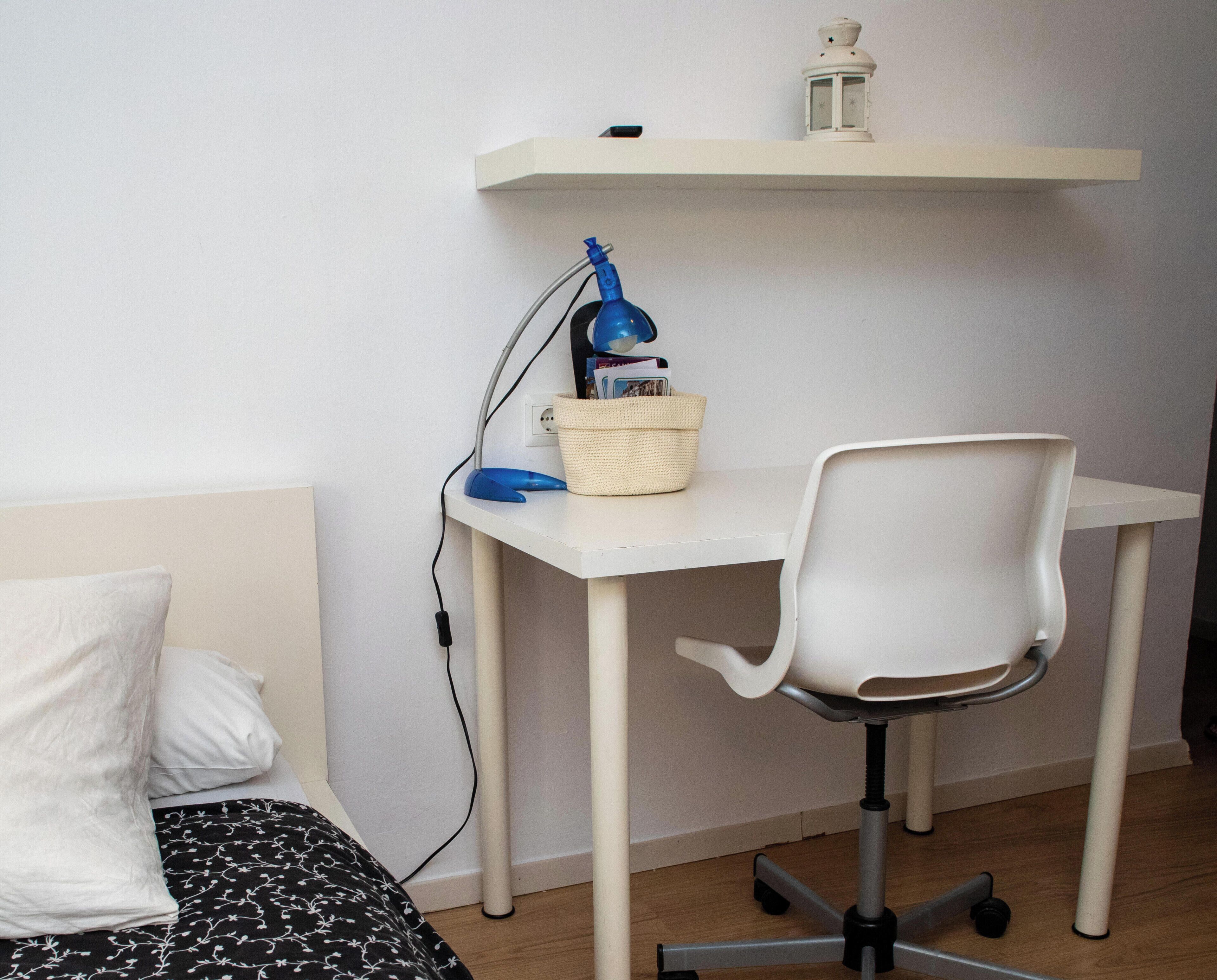 A small room with a bed, a desk with a chair, a lamp, a basket, and a wall-mounted shelf with a lantern.