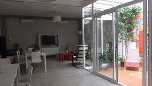 Un'ampia stanza con un'ampia porta-finestra scorrevole che dà su uno spazio esterno, una TV a schermo piatto montata a parete e diversi tavoli e sedie bianche disposti per mangiare o riunioni.