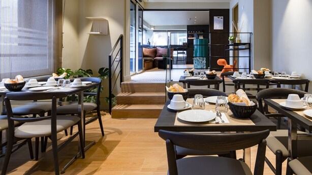 Een moderne eetruimte met gedekte tafels voor het ontbijt, een trap naar een woonruimte, en een uitzicht op de stad door grote ramen.