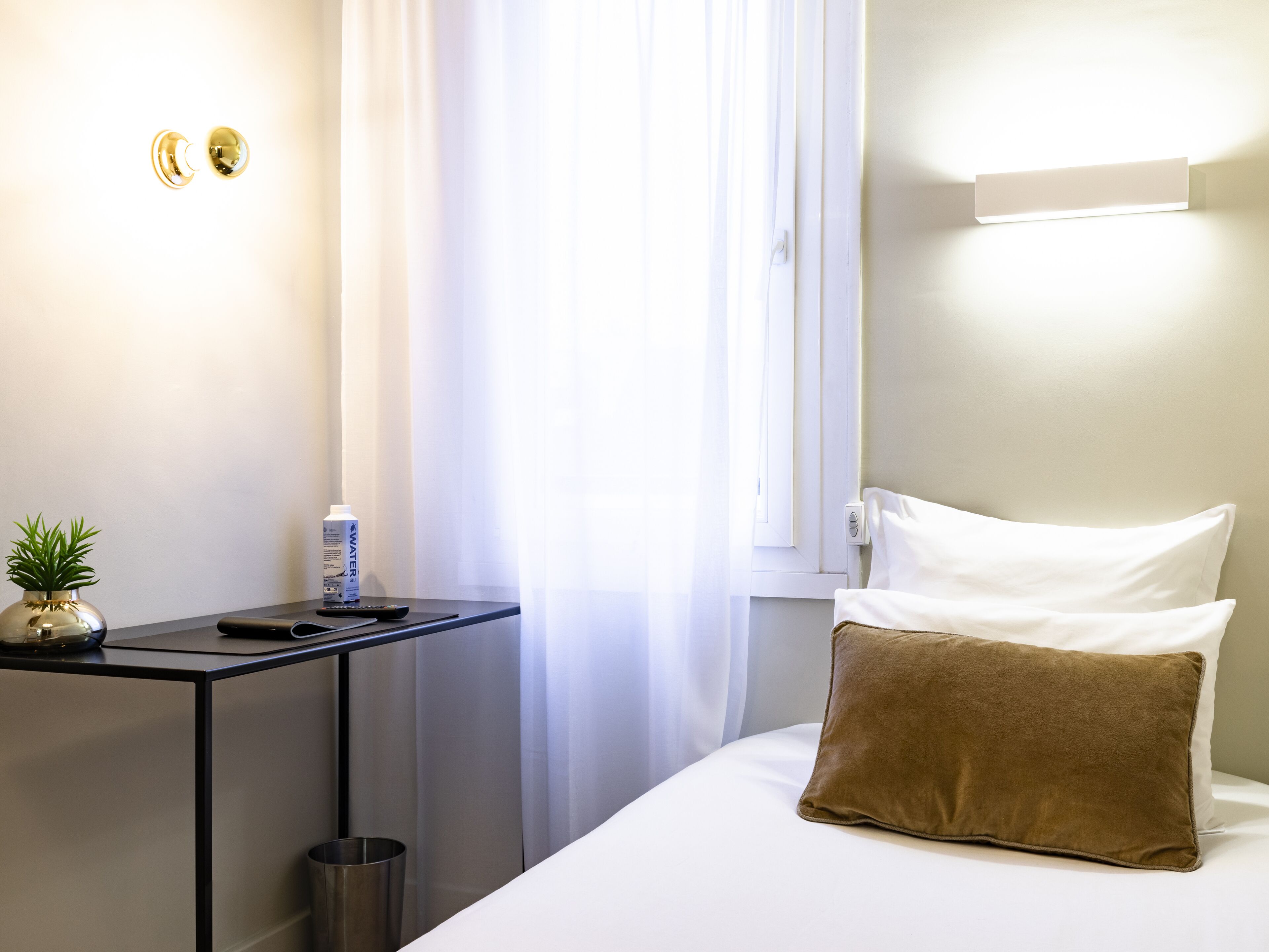 A hotel room with a bed, a bedside table with a remote control, a water bottle, and a small plant.