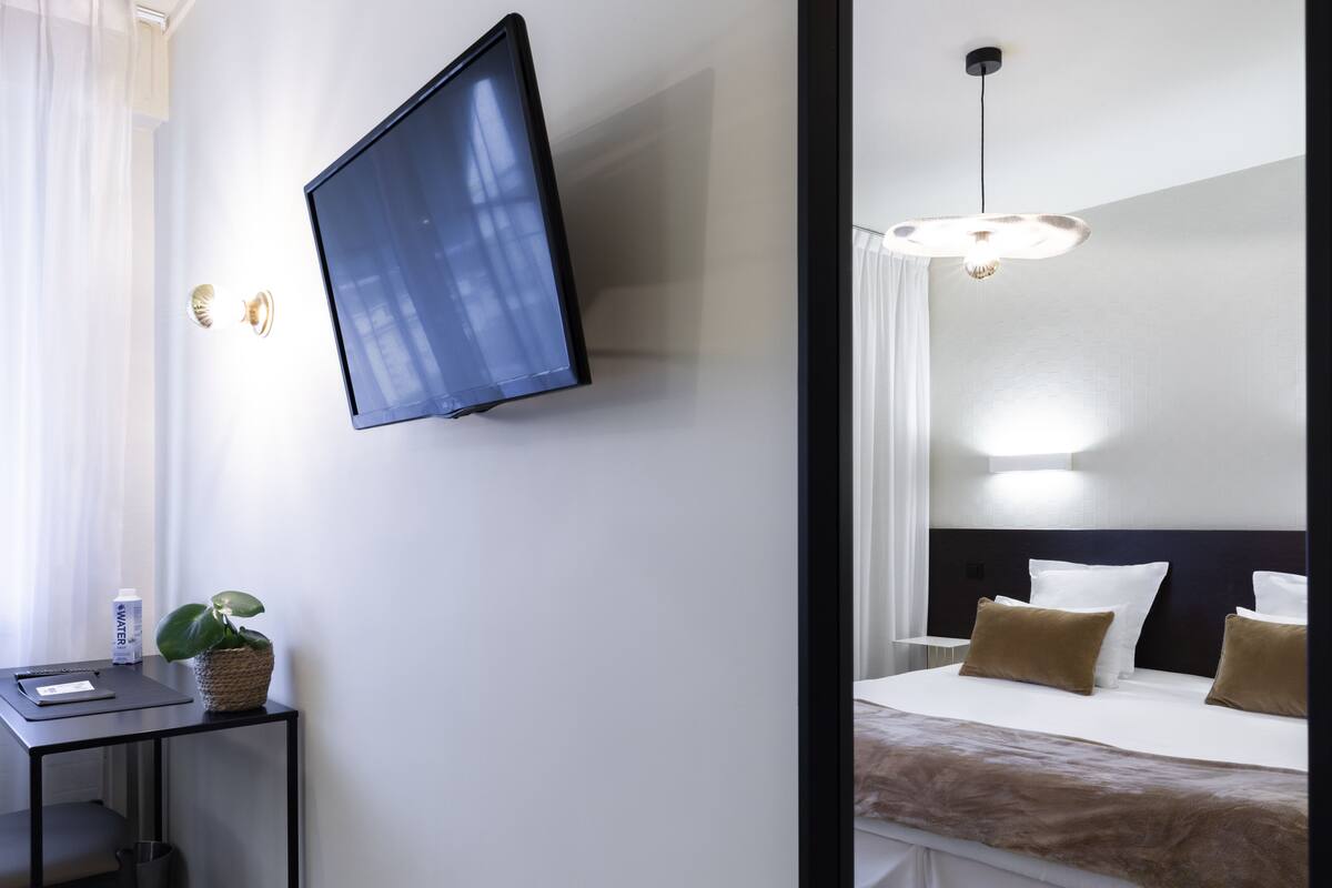 Habitación de hotel con televisor de pantalla plana en la pared, un escritorio con planta y una cama con ropa de cama blanca y marrón.