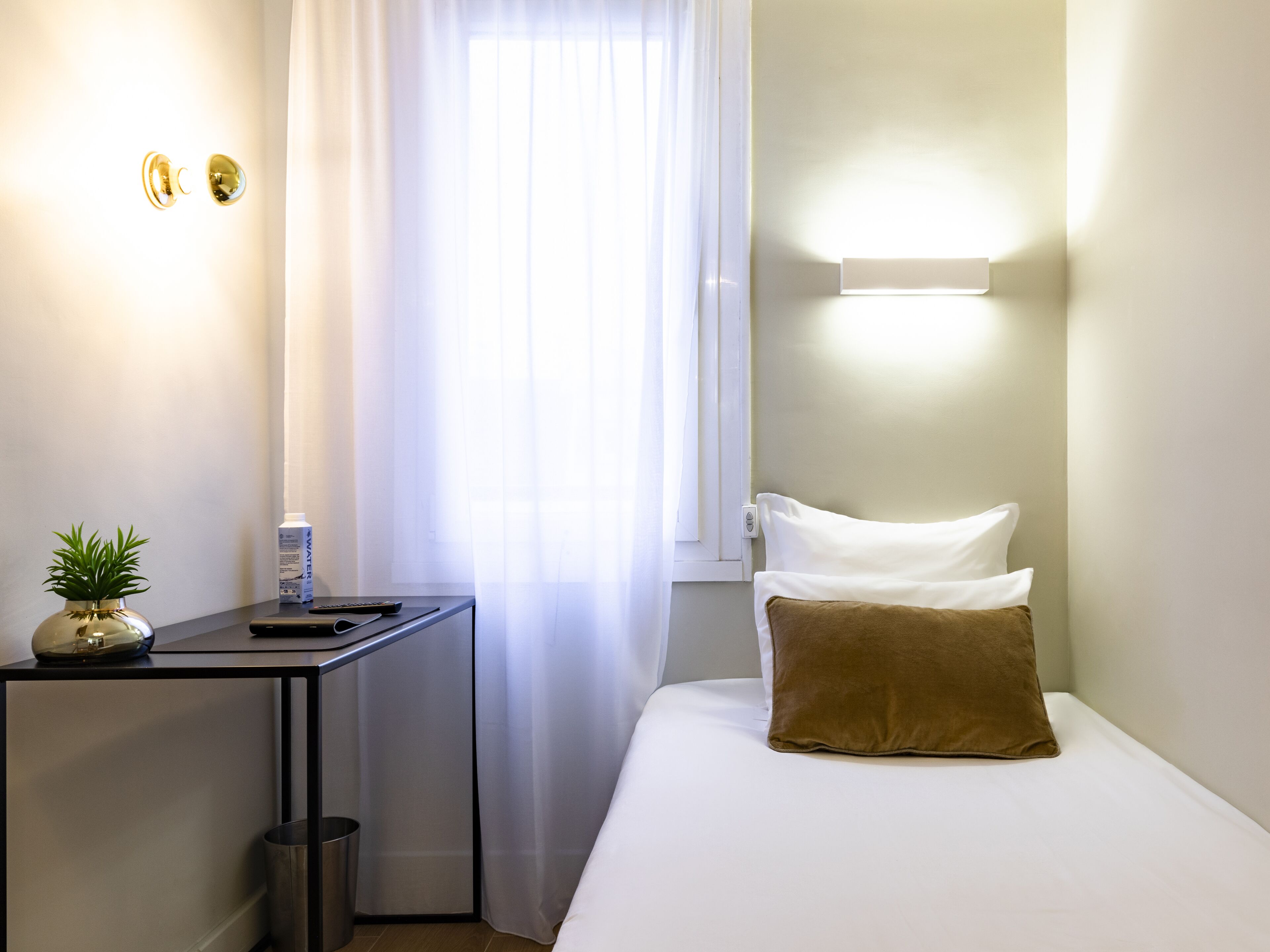 A hotel room with a bed, a bedside table, a small potted plant, a remote control, and a bottle of water.