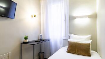 Habitación de hotel con una cama, un televisor montado en la pared, una mesita con una planta y una botella, una ventana con cortinas y un radiador.