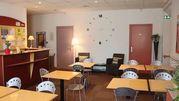 A hotel lobby with a reception desk, multiple tables and chairs, a clock on the wall, and a few potted plants.
