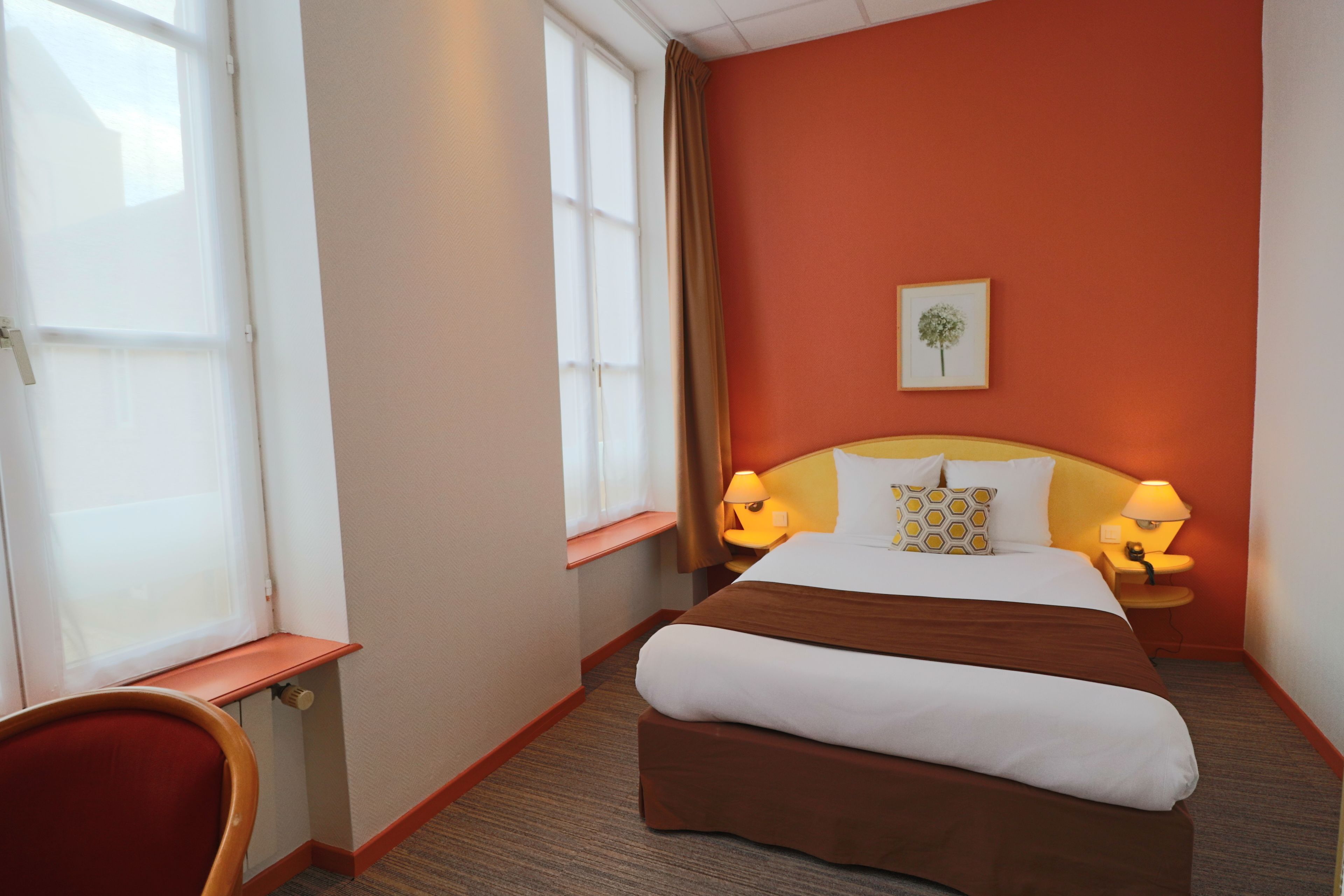 Une chambre d’hôtel avec un grand lit, deux lampes de chevet, une fenêtre avec des rideaux, une petite table et un tableau encadré au mur.