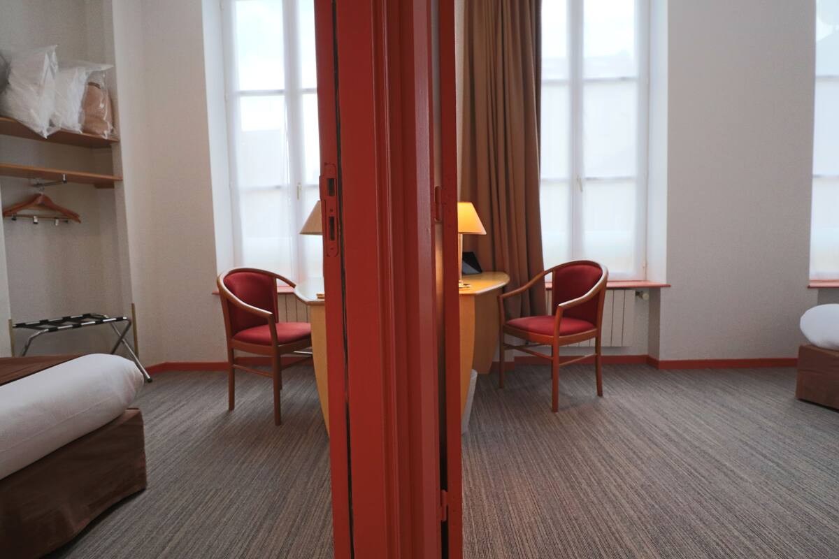 A hotel room with a bed, two red chairs, a small table with a lamp, and a window with curtains.